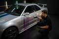 Technician spraying iron fallout remover on the lower panel of a silver 2001 Nissan Skyline GT-R R34 with blue racing stripes, showing purple-red chemical reaction on the paint in a dark luxury detailing studio
